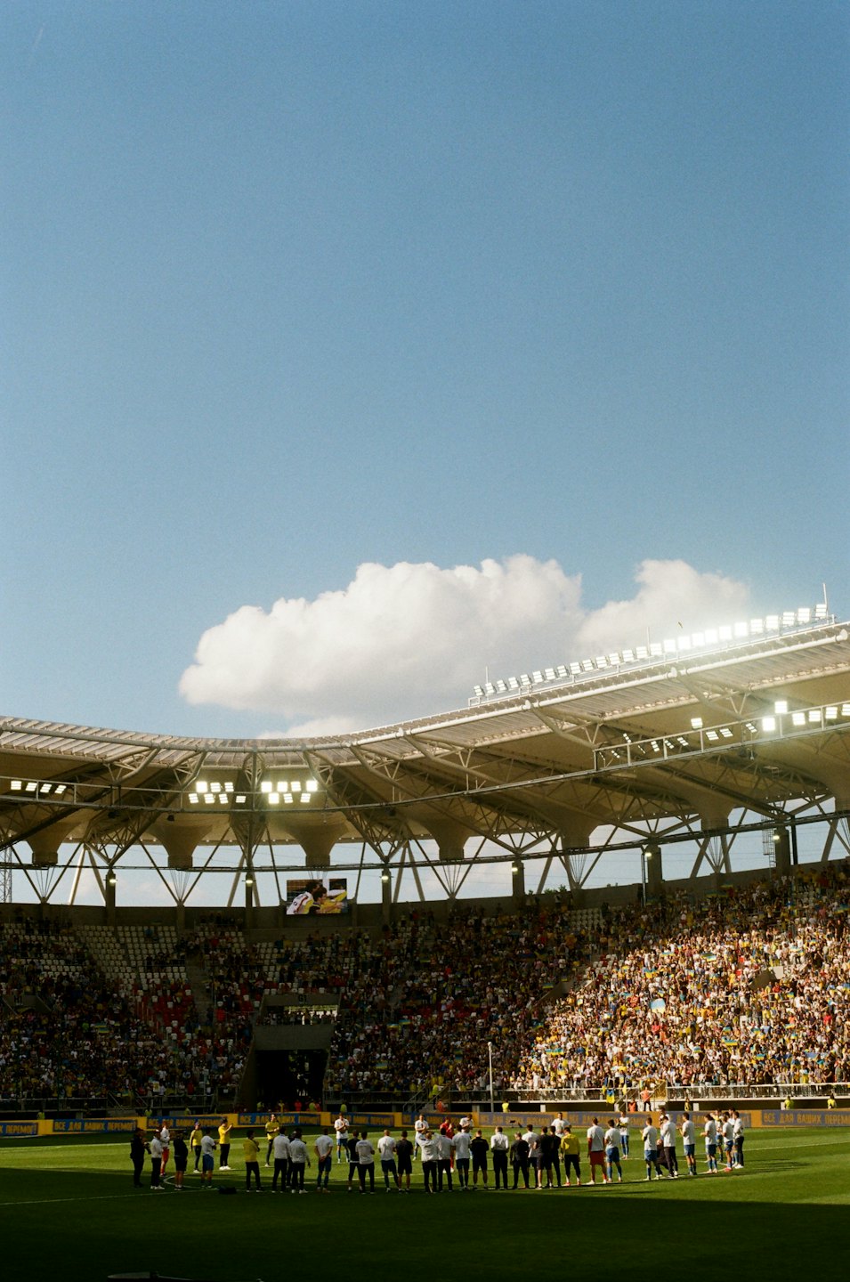 Athletes on a sunny sports field facing a packed stadium.